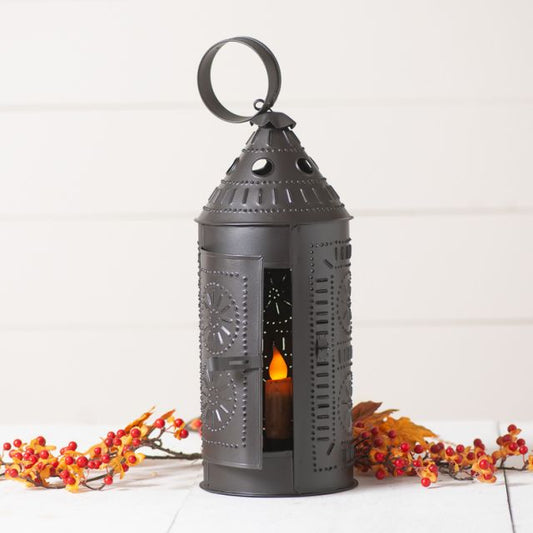 A smokey black finished metal Sturbridge Lantern with a chisel tin punch design, featuring a battery operated taper candle lit inside, placed on a white surface with red and yellow leaves around it.