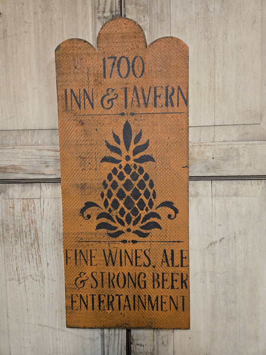 A rustic wooden sign for '1700 Inn & Tavern' featuring a black stenciled pineapple design and text advertising fine wines, ale, strong beer, and entertainment, shown against a weathered light wood plank background.