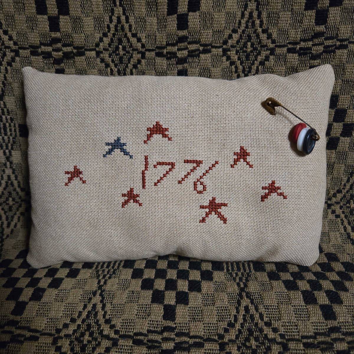 A square linen pillow with embroidered red and blue stars and the year '1776' in red thread, resting on a patterned black and beige blanket. A small safety pin with a striped ball charm is pinned to the top right corner of the pillow.