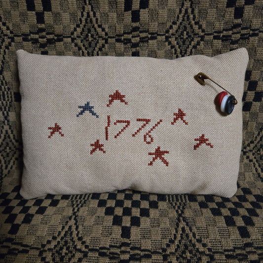 A square linen pillow with embroidered red and blue stars and the year '1776' in red thread, resting on a patterned black and beige blanket. A small safety pin with a striped ball charm is pinned to the top right corner of the pillow.
