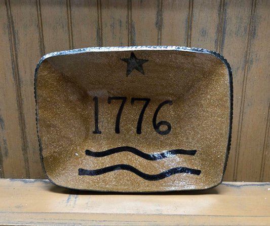 A rustic rectangular ceramic tray with rounded corners, featuring a distressed gold-brown glaze and black hand-painted details including a star, the year '1776', and two wavy lines, set against a wooden background.