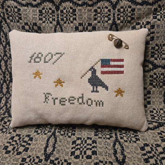Decorative pillow with cross-stitched design featuring an American flag, airplane, and the word 'Freedom' on a textured fabric background.