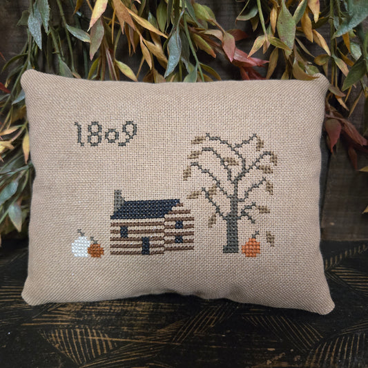 Decorative pillow with cross-stitched design of a house and tree on a wooden surface.