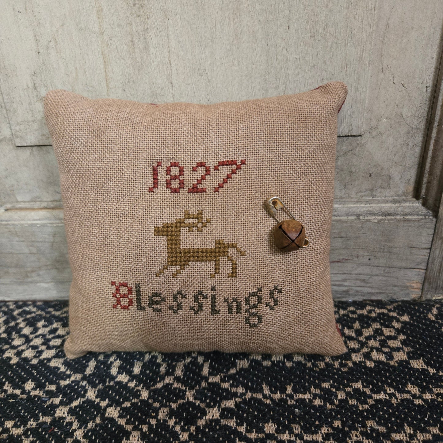 A square cross stitch pillow with the text '1827 Blessings' and a reindeer motif, displayed against a wooden background.