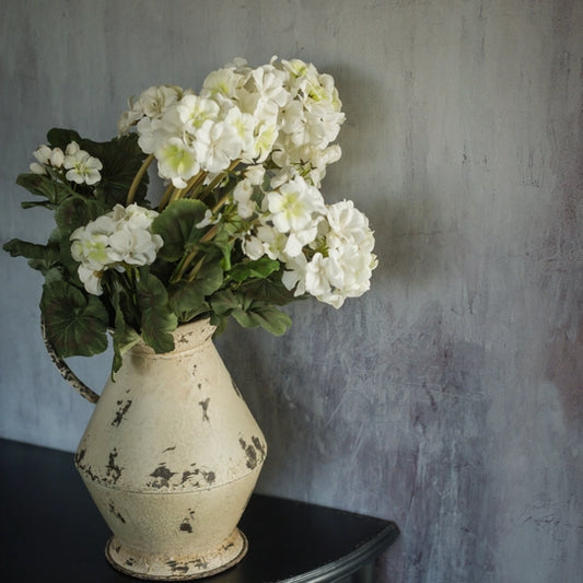 An artificial white geranium bush in a vintage style beige vase, placed on a surface against a gray background.