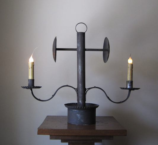 A metal table lamp with two arms, each holding a candle, set on a wooden table against a textured wall.