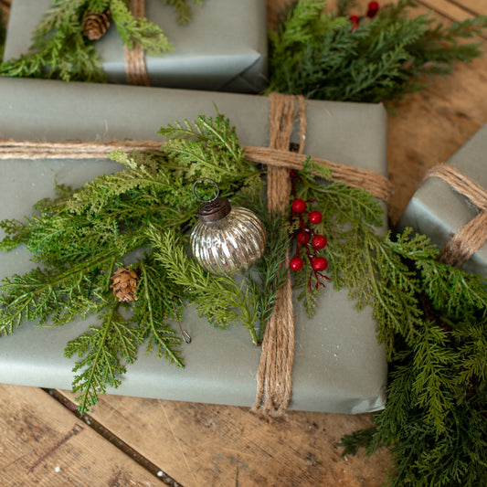 A silver ribbed kugel ornament with red berries, attached to a green wreath and a brown paper gift bag on a wooden surface.