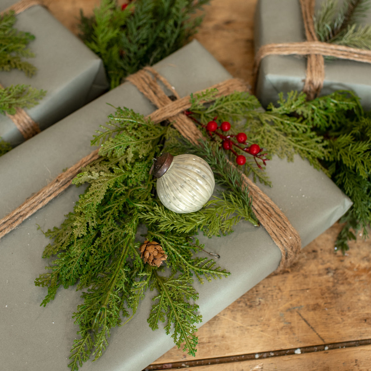 A wrapped gift box in matte gray paper, tied with natural jute twine and decorated with green pine branches, red berries, a small pinecone, and a ribbed white ornament, set on a rustic wooden surface.