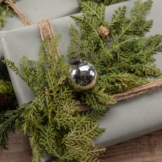 A 2-inch silver kugel ornament placed on a bed of green pine needles, with a burlap bow on a gift wrapped in a neutral-colored paper.