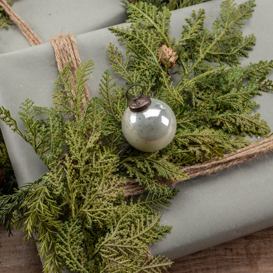 A 2-inch white glass Kugel ornament with green artificial foliage attached, placed on a textured surface with a rope.