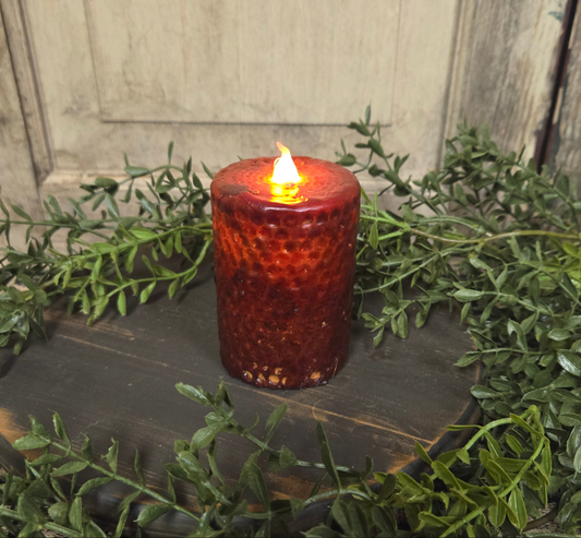 A burgundy honeycomb pillar candle with a lit flame, placed on a dark surface surrounded by green foliage.