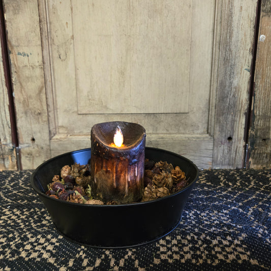 A black moving flame pillar candle on a rustic metal dish with a flickering light, placed on a patterned mat against a wooden background.