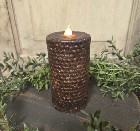 A black honeycomb patterned pillar candle with a lit flame on top, placed on a wooden surface with greenery in the background.