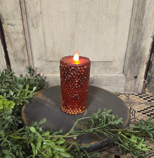 A battery-operated burgundy honeycomb-patterned LED pillar candle displaying a flame-shaped light, placed on a circular dark base surrounded by greenery.