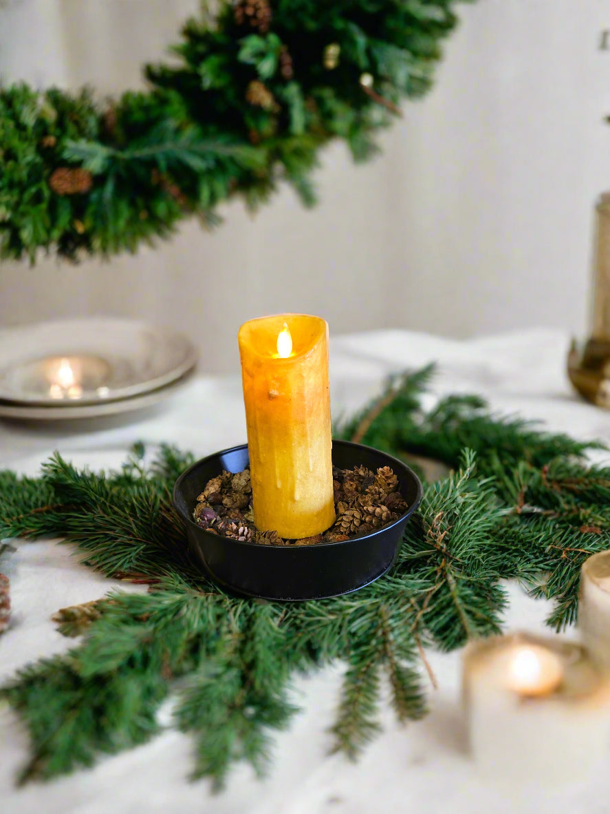 A flameless, moving flame pillar candle in a burnt mustard color, displayed on a dark surface with a patterned texture, against a wooden background.