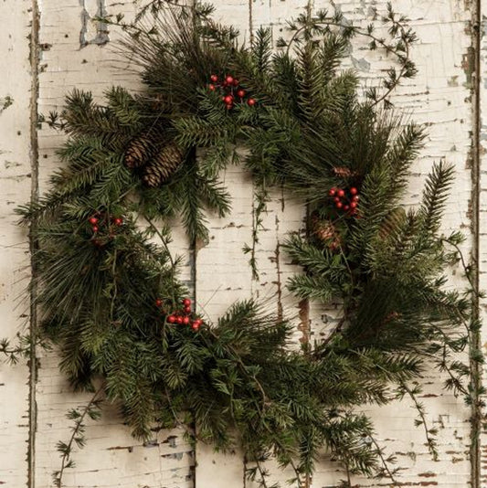 A round wreath made of mixed pine branches with red berries, displayed on a white wooden background.