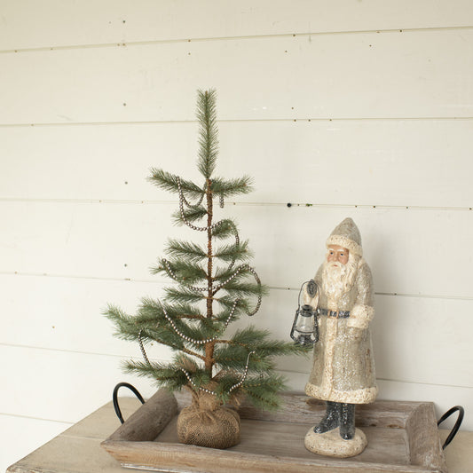 An artificial foxtail pine tree with a rustic burlap base, placed on a wooden surface against a white wall, decorated with ornaments and a figure of Santa Claus.
