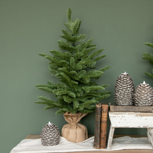 Artificial pine tree decoration with tied burlap base, displayed on a white surface next to a wooden crate with pine cones.