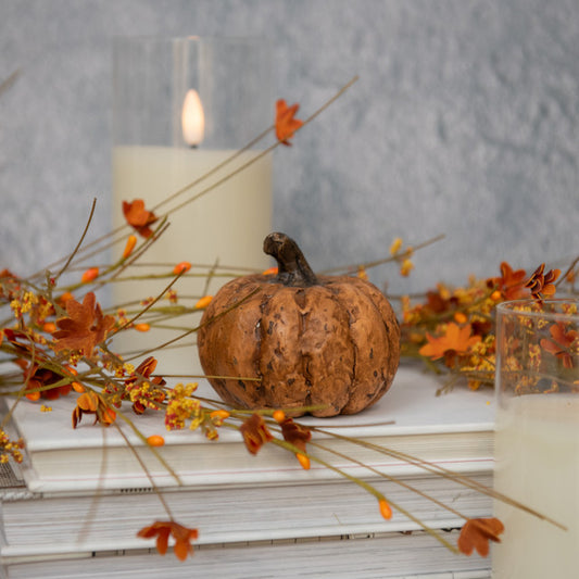 A terracotta sculpture of a short pumpkin with a textured surface, placed on a book, surrounded by dried flowers and a candle.