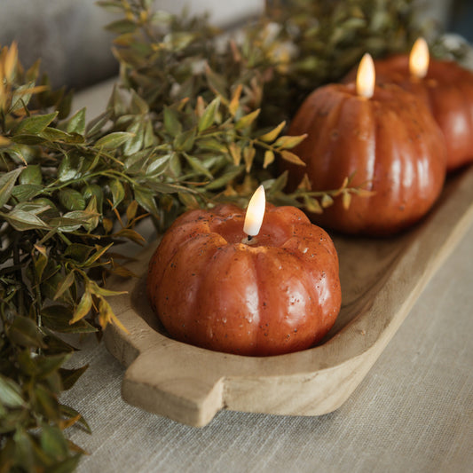 Decorative candles shaped like pumpkins with a lit candle on a wooden tray with greenery.