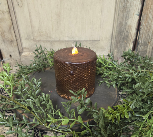 A black honeycomb-patterned pillar candle with a lit flame on top, surrounded by greenery and placed on a black round holder.