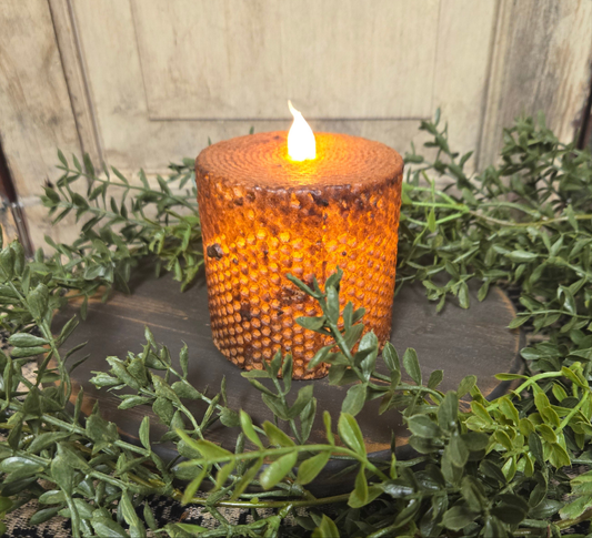 A brown honeycomb pillar candle with a lit flame on top, surrounded by green foliage and placed on a dark circular holder.