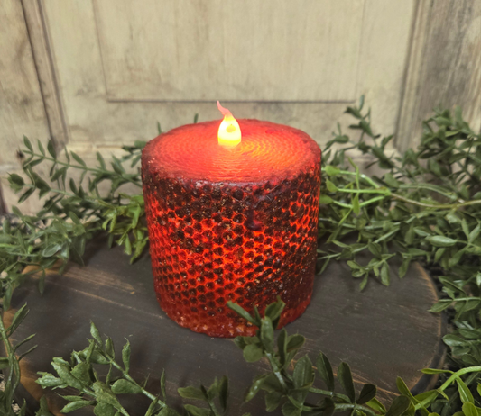 A burgundy wrapped honeycomb LED pillar candle with a lit flame, placed on a wooden surface with greenery around it.