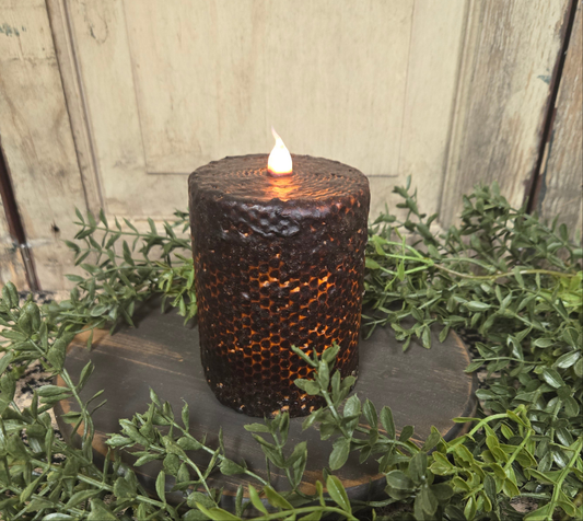 A black honeycomb patterned pillar candle with a lit flame on top, placed on a wooden holder with greenery around it.