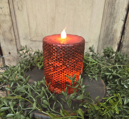 A burgundy wrapped honeycomb LED pillar candle with a flame-shaped lightbulb flickering, placed on a green mossy surface.