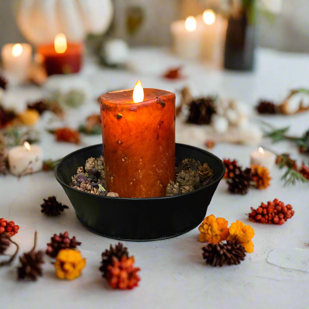 A 3 inch by 4 inch moving flame pillar candle in burnt orange color, placed on a black dish on a lace doily with a wooden background.