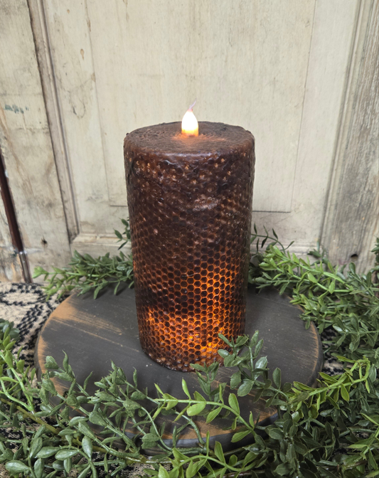 A black honeycomb-patterned pillar candle with a lit flame on top, placed on a round brown base, surrounded by greenery.