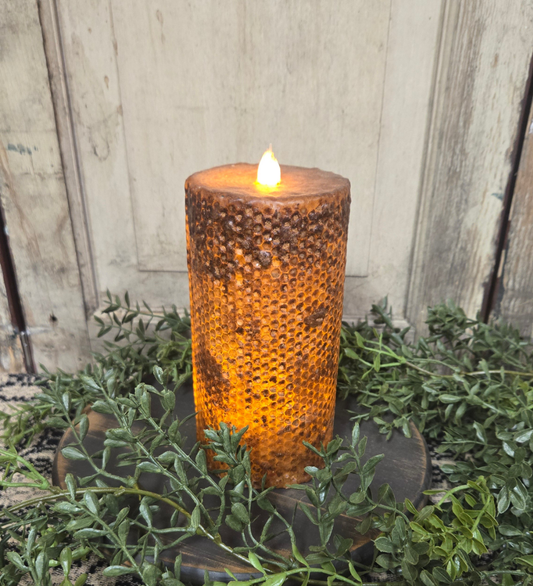 A brown honeycomb pillar candle with a lit flame on top, placed on a wooden stand surrounded by greenery.