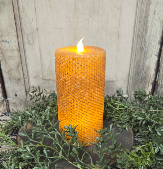 A mustard yellow honeycomb LED pillar candle placed on greenery, with a black honeycomb-patterned wax coating and a flame-shaped light at the top.