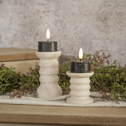 Two black tealight candles with 3D flame sitting on white pedestals with greenery in background.