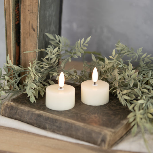 Two cream-colored tealight candles with a 3D flame effect, placed on an open book, surrounded by green foliage.