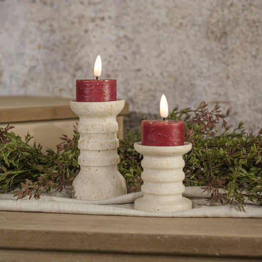 Two red tealight candles placed in white 3D flame holders, surrounded by greenery on a wooden surface.