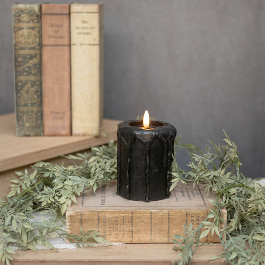A black 3D flame pillar candle with an on/off button, placed on a wooden surface with books and decorative foliage in the background.