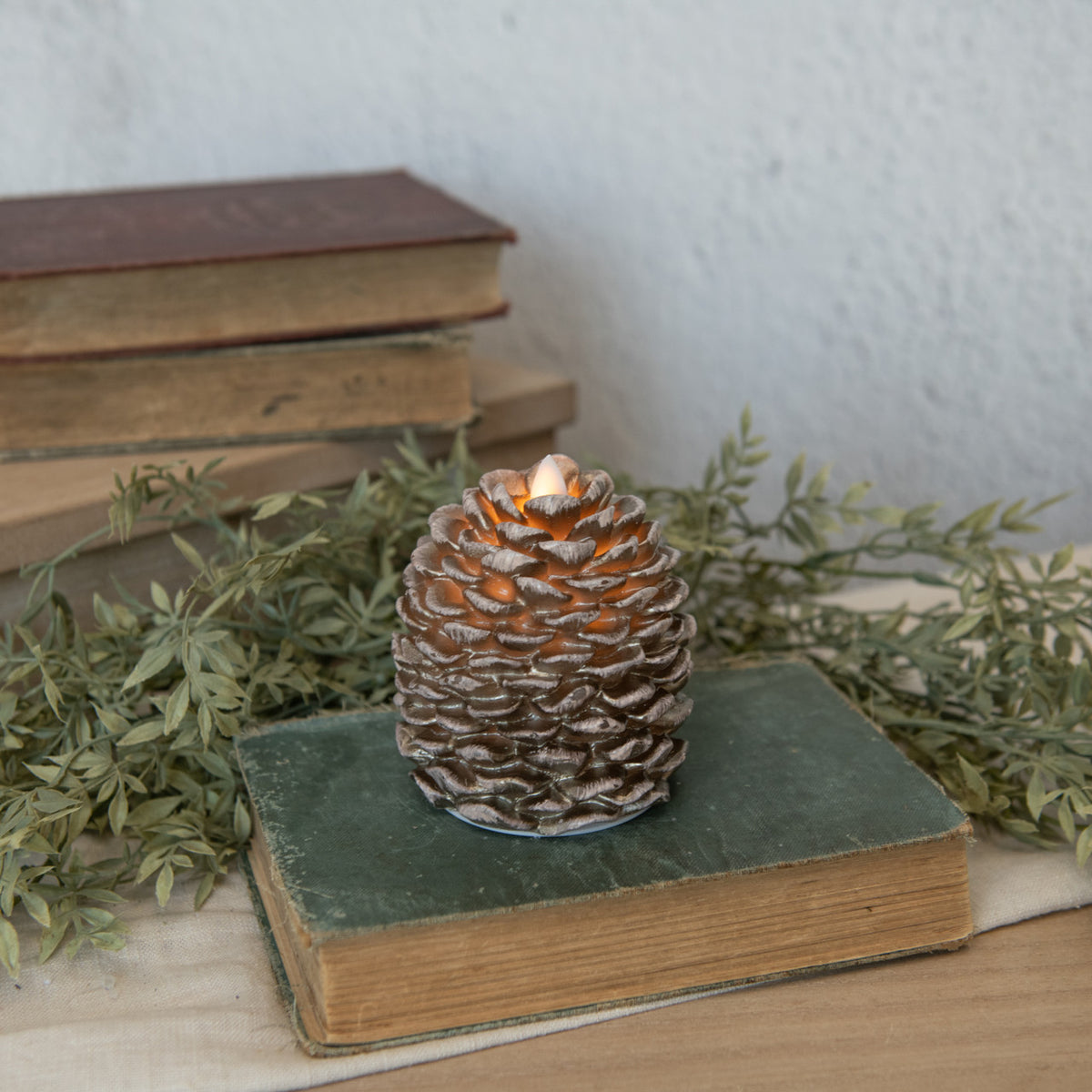 A pinecone-shaped candle with a realistic moving flame, placed on an open book, surrounded by greenery and an old-fashioned lamp.