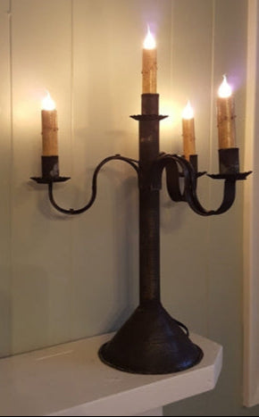 A black metal 4 light candelabra tin table light with lit candles, placed on a white surface.