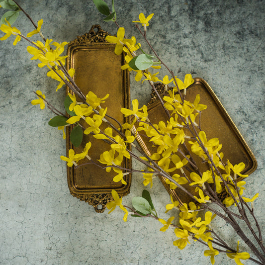 A decorative metal object with a gold finish, resembling a stem, adorned with artificial yellow forsythia flowers against a concrete background.