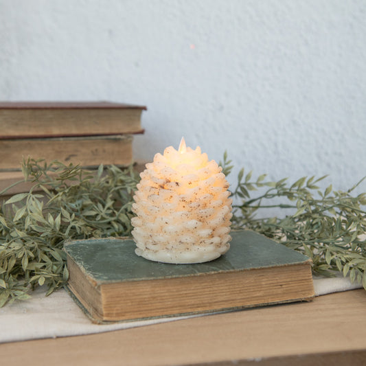 A cream pinecone-shaped candle with a moving flame, placed on an open book, with a background of books and decorative foliage.