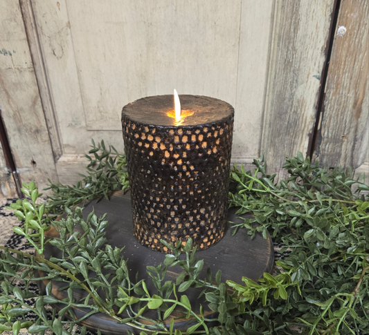 A black beeswax honeycomb pillar candle with a realistic moving flame, placed on a round black base, surrounded by green foliage.