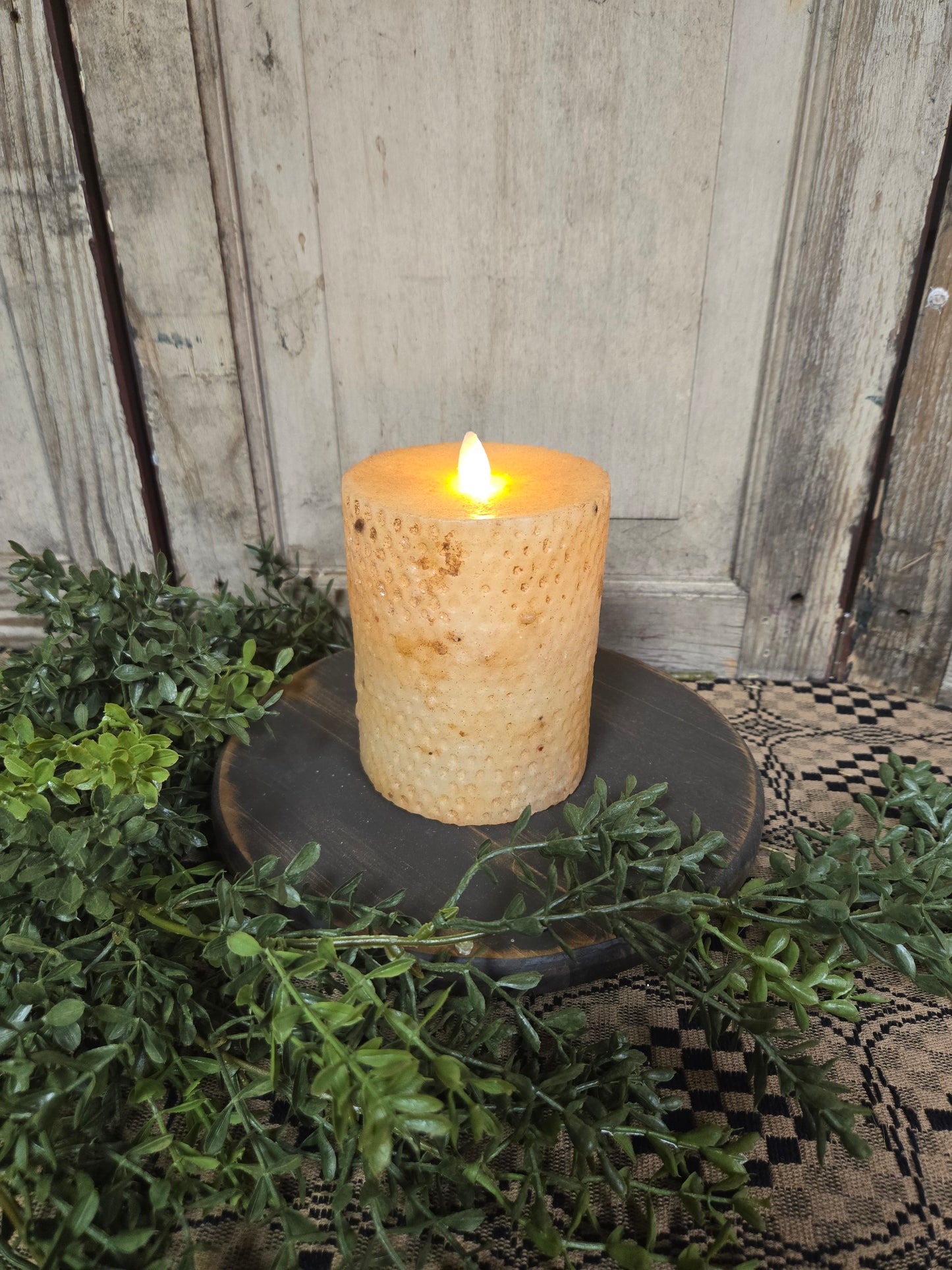 A 5-inch beeswax honeycomb pillar candle with a realistic moving flame, placed on a dark candle holder, surrounded by greenery.