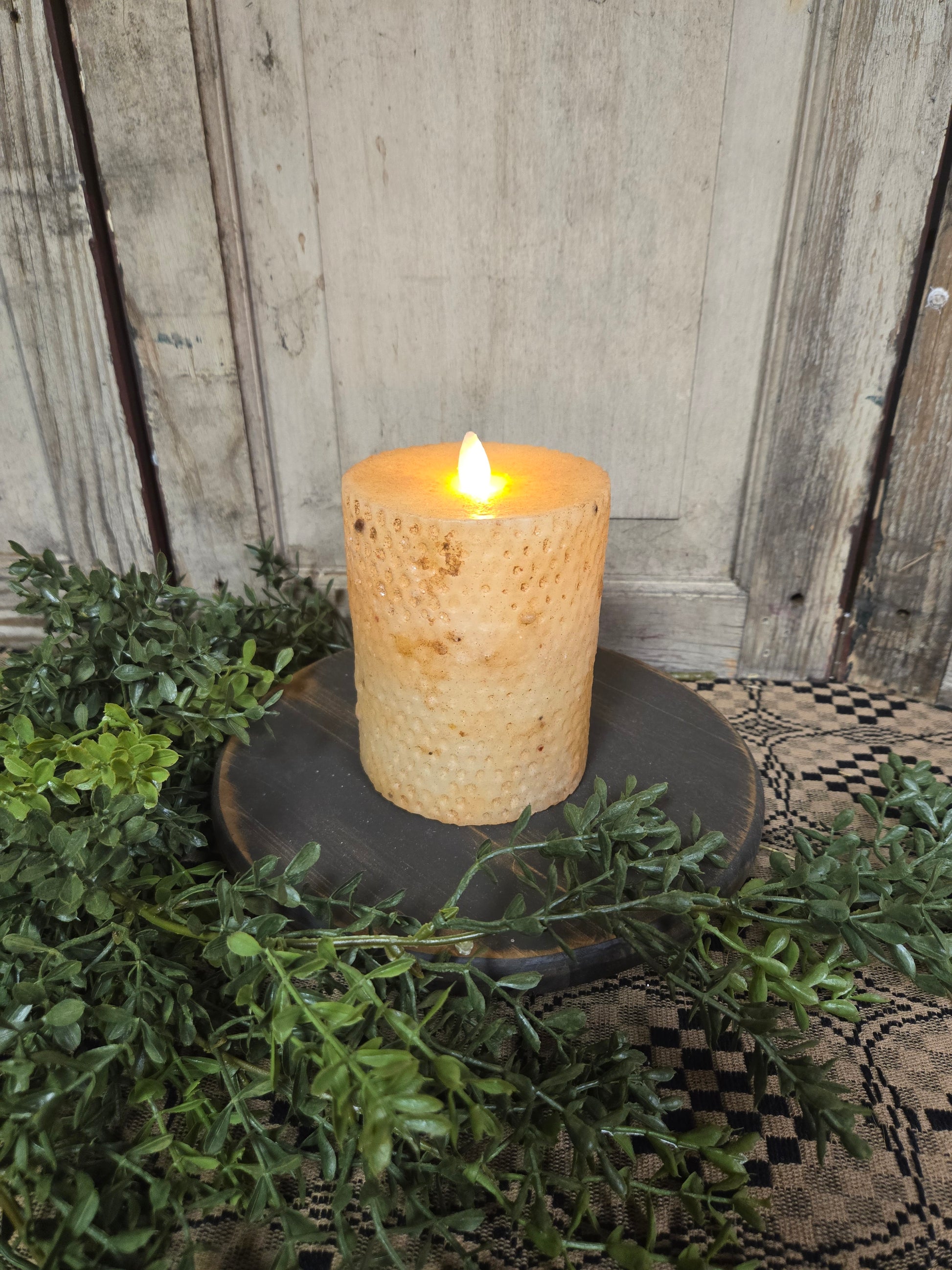 A 5-inch beeswax honeycomb pillar candle with a realistic moving flame, placed on a dark candle holder, surrounded by greenery.