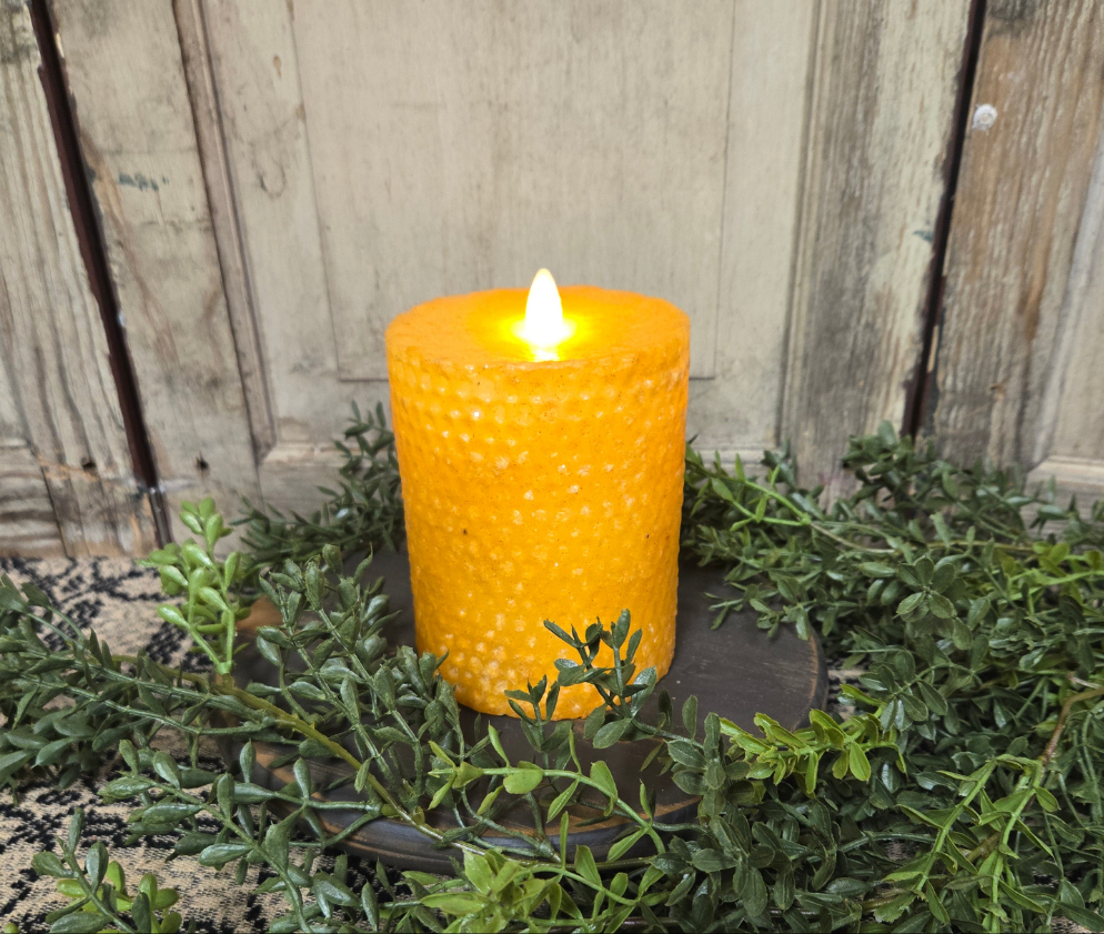 A battery-operated beeswax honeycomb pillar candle with a realistic moving flame, placed on a darker surface with greenery around it.