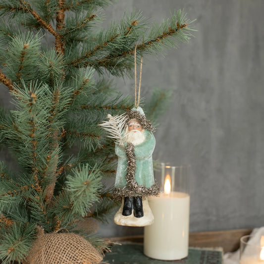 A small hanging Christmas ornament shaped like a Santa figure in a pale green coat with silver tinsel trim, displayed on a pine tree branch beside a lit white candle against a soft gray backdrop.
