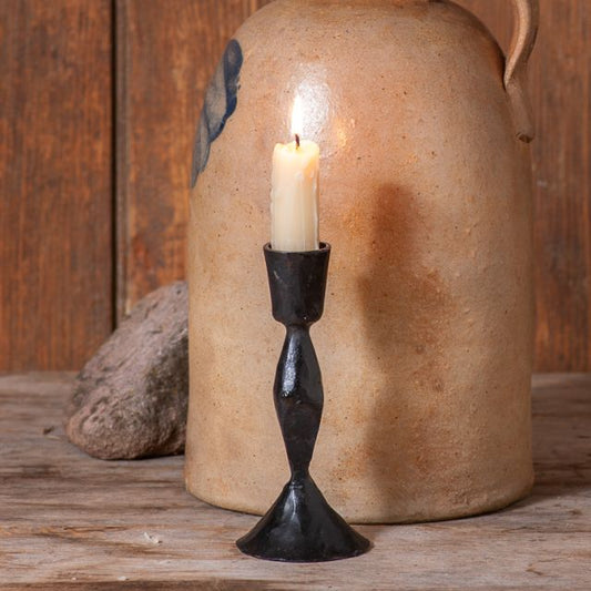 A black cast iron taper candle holder with a lit candle, placed on a wooden surface next to a ceramic jug.