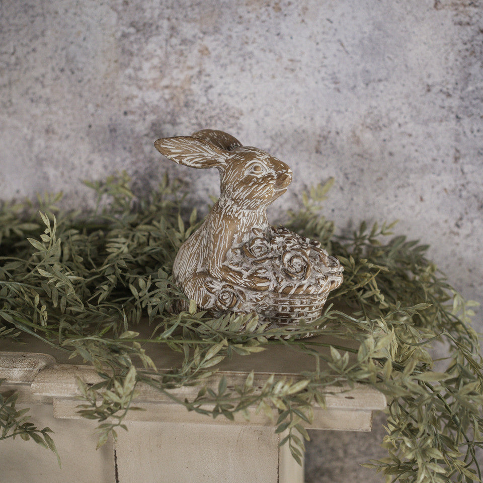 A resin figurine of a beige bunny with a full basket, placed on a surface with artificial greenery in the background.