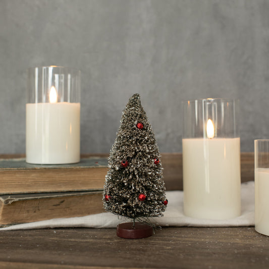 A 6-inch green bottlebrush tree with a realistic appearance, placed on a white surface next to three lit candles in clear glass holders.