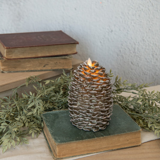 A pinecone-shaped candle with a realistic flame, placed on a book, with a background of books and decorative foliage.